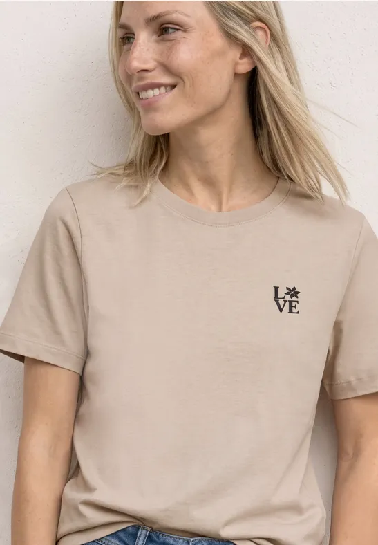 Vrouw lacht, draagt beige T-shirt met klein 'LOVE'-logo, tegen een lichte achtergrond.