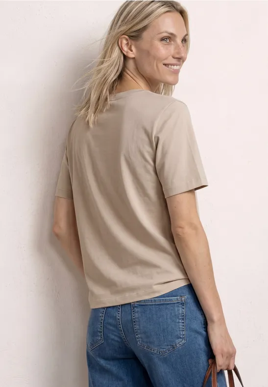 Achteraanzicht van persoon met beige T-shirt, blauwe spijkerbroek, blond haar en klein tasje.