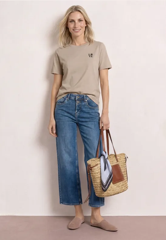 Vrouw tegen lichte achtergrond met beige T-shirt, wijde cropped jeans, rieten tas en platte schoenen.