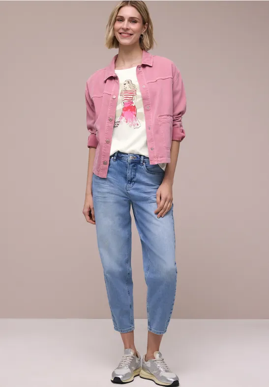 Modell trägt pinke Jeansjacke, bedrucktes weißes T-Shirt, lockere blaue Jeans und Sneaker vor neutralem Hintergrund.