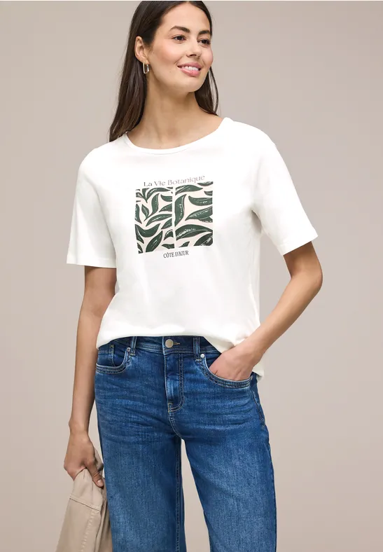Eine Frau trägt ein weißes T-Shirt mit grünem Muster und blaue Jeans.