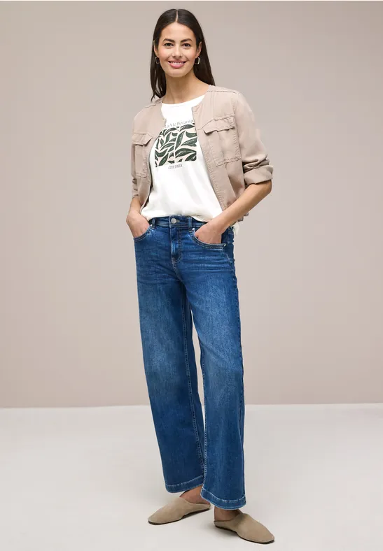 Frau steht vor neutralem Hintergrund, trägt helle Jacke, weißes T-Shirt mit grünem Print, weite Jeans und flache Schuhe.