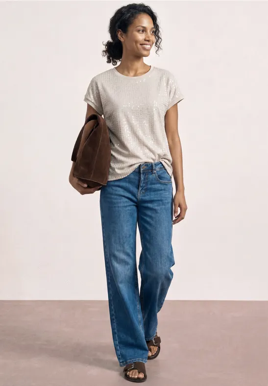 Persoon in wit t-shirt, blauwe jeans en sandalen, met een bruine jas onder de arm.