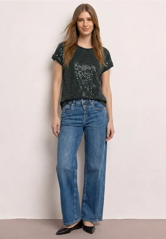 Vrouw in een zwart bedrukte t-shirt, rechte blauwe jeans en zwarte schoenen tegen een lichte achtergrond.
