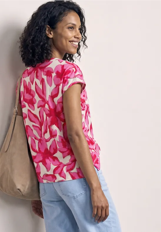 Femme de profil en chemise rose à fleurs, jean clair et grand sac beige, devant un fond neutre.