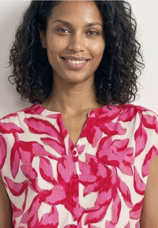 Femme aux cheveux bouclés porte un chemisier rose et rouge à motif abstrait sur fond blanc.