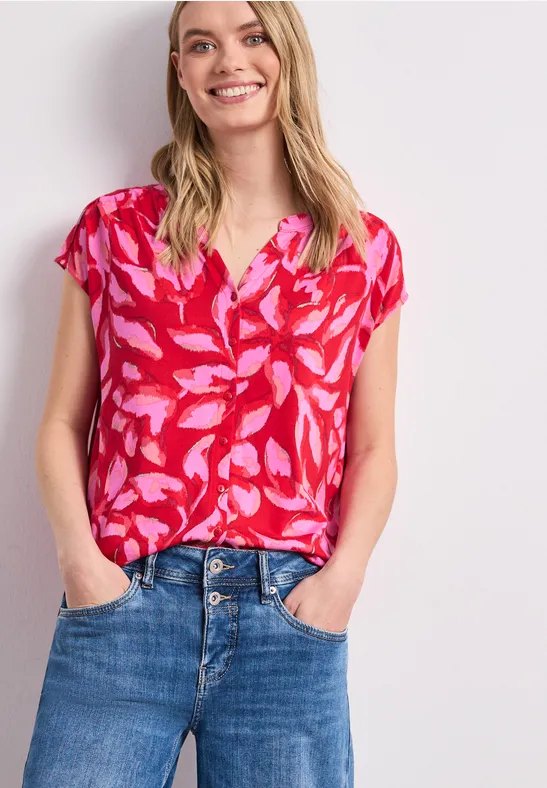 Vrouw draagt rood bloesje met roze bladmotief en een blauwe spijkerbroek.