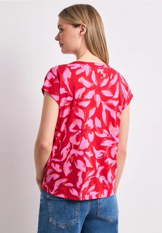 Vrouw van achteren in een rood shirt met roze bladprint en een blauwe spijkerbroek.