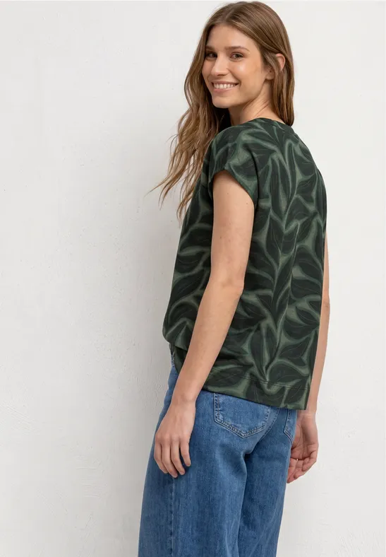 Femme vue de dos portant un chemisier vert à motif feuilles et un jean bleu.