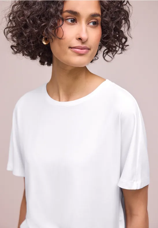 Eine Person mit lockigem Haar und goldenen Creolen trägt ein schlichtes weißes T-Shirt.