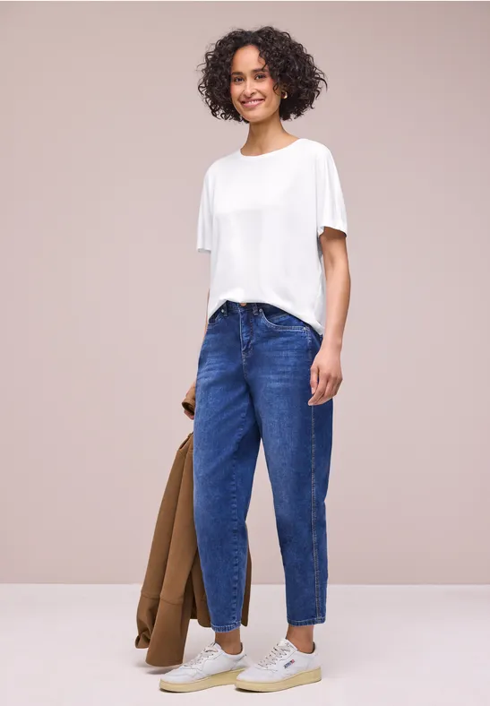 Person trägt weißes T-Shirt, blaue Jeans und hält eine braune Jacke vor hellem Hintergrund.