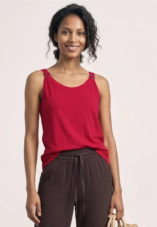Een vrouw draagt een rood mouwloos topje en losse bruine broek met koord, ze houdt een voorwerp in haar hand.