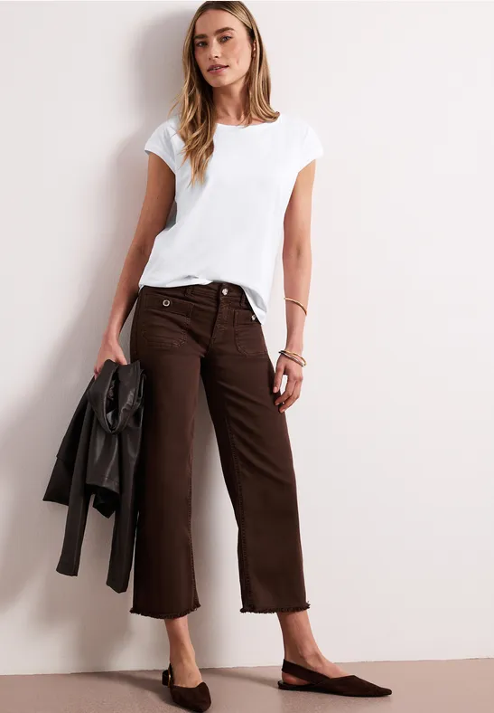 Vrouw in wit T-shirt en bruine culotte, met zwarte jas in hand, tegen een lichte muur.