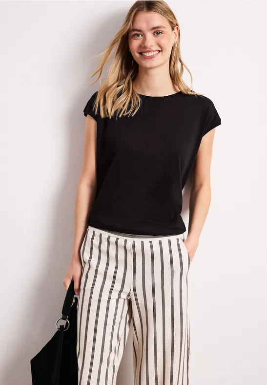 Vrouw in zwart t-shirt en wit gestreept broek, handen in zakken, staand voor een witte achtergrond.