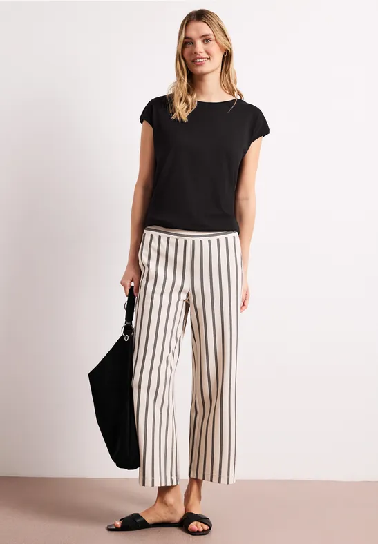 Vrouw in studio met zwarte korte mouw top, brede gestreepte broek, sandalen en zwarte tas.