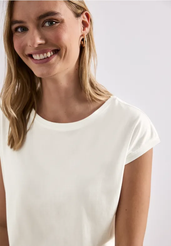 Junge Frau in weißem T-Shirt lächelt, trägt Ohrstecker, helle Studioaufnahme, schulterlange blonde Haare.