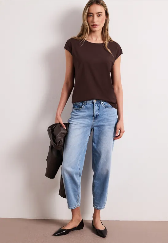 Vrouw met bruin T-shirt, losse blauwe spijkerbroek, zwarte ballerina’s, houdt jas in hand.