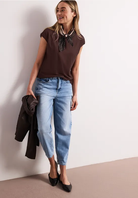 Vrouw in bruin shirt, sjaal, lichtblauwe cropped jeans, zwarte ballerina's, met jas in hand.