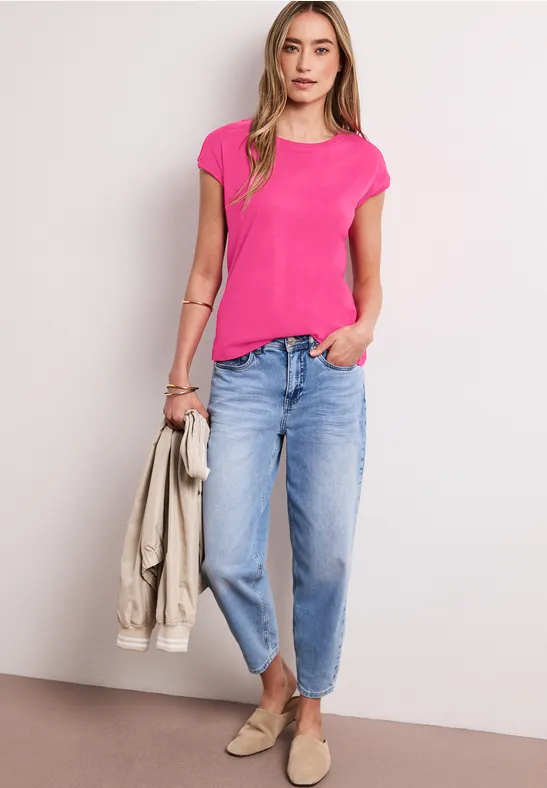 Eine Frau trägt pinkes T-Shirt, hellblaue Jeans, hält helle Jacke und trägt beige Schuhe.