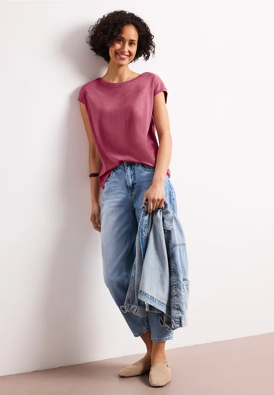 Frau steht in rosa Top und weiten Jeans, hält hellblaue Jeansjacke in der Hand.