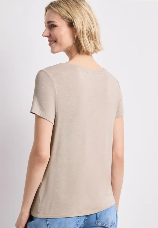 Vue de dos d’une femme aux cheveux blonds courts portant un t-shirt beige et un jean bleu sur fond blanc.