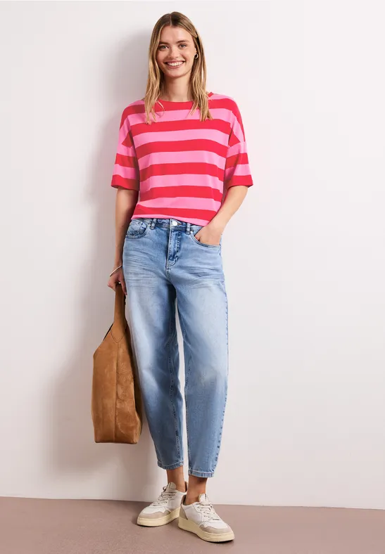 Eine Frau trägt ein pink-rot gestreiftes T-Shirt, weite hellblaue Jeans, weiße Sneaker und hält eine braune Tasche.