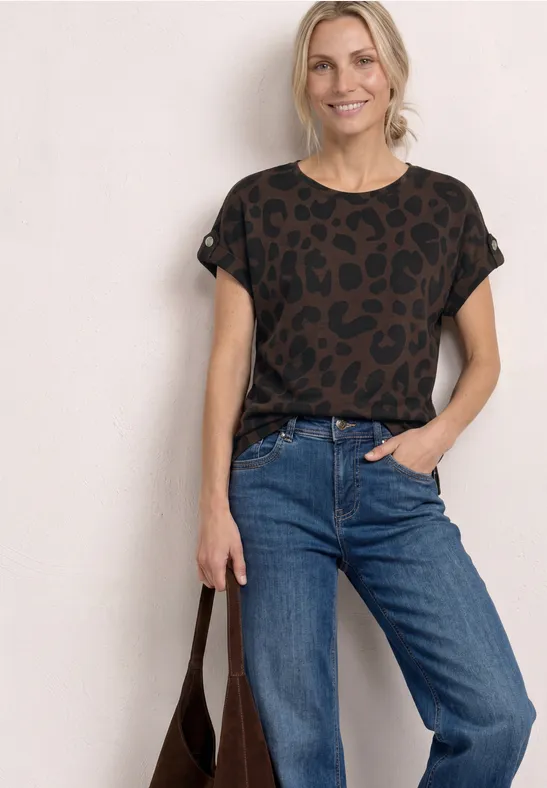 Vrouw in casual outfit: luipaardshirt, blauwe jeans en bruine tas tegen witte achtergrond.