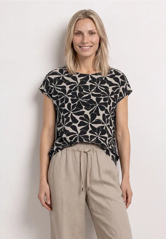 Vrouw in zwarte t-shirt met grote witte bloemenprint en beige, losse broek voor witte achtergrond