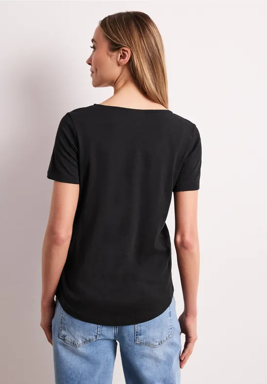 Frau von hinten trägt schwarzes T-Shirt und blaue Jeans vor hellem Hintergrund.