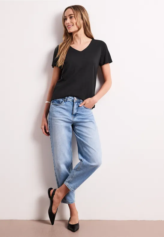 Eine Frau steht lässig in einem schwarzen T-Shirt und hellblauen Jeans vor hellem Hintergrund.