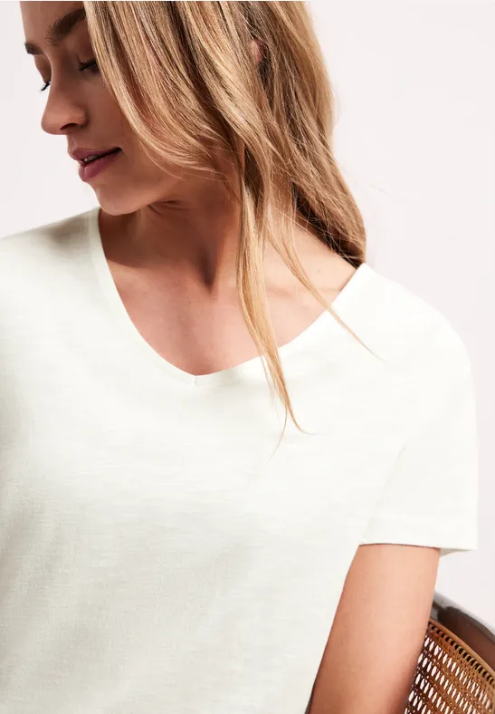 Frau in schlichtem weißen V-Shirt, seitlich mit blondem Haar posierend vor hellem Hintergrund.