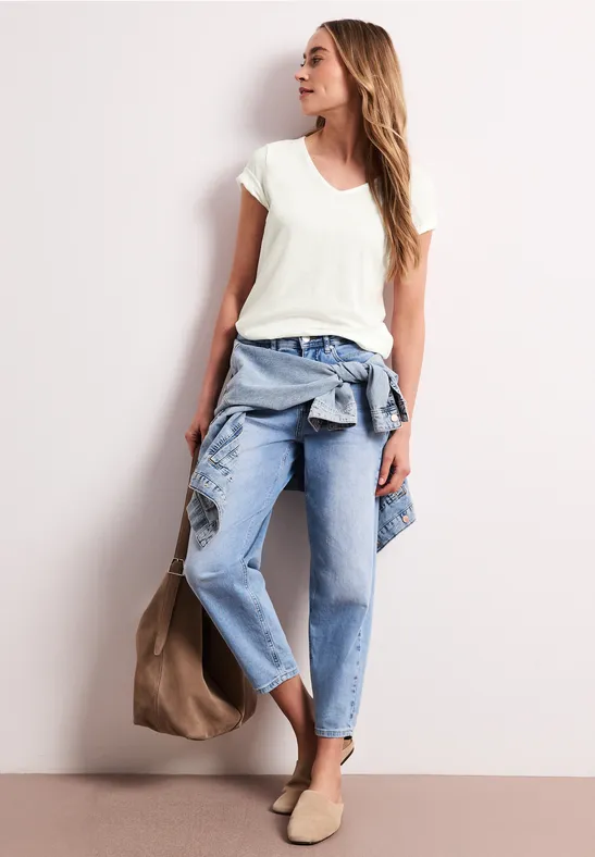 Frau in legerem Outfit mit weißem T-Shirt, blauen Jeans, Jacke um die Taille, beiger Tasche und Slippern