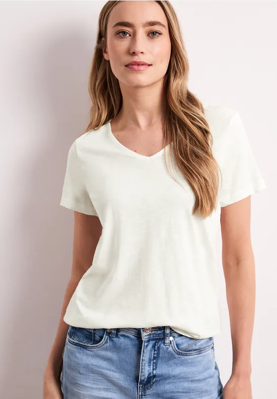 Frau trägt weißes T-Shirt und blaue Jeans vor hellem Hintergrund.