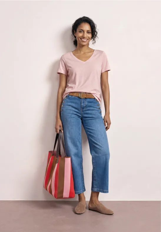 Vrouw in roze T-shirt, blauwe spijkerbroek en beige espadrilles met een gestreepte tas voor een lichte achtergrond.