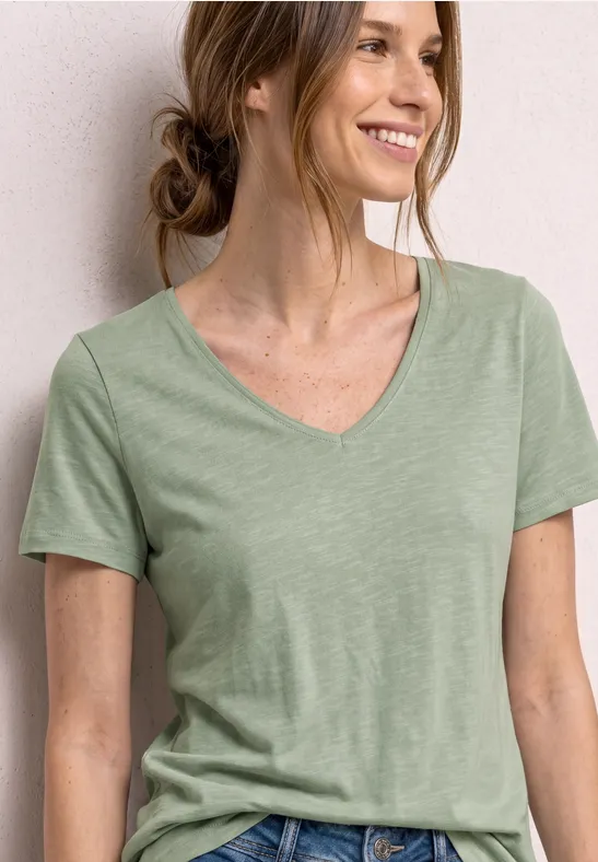 Frau in hellgrünem V-Ausschnitt-T-Shirt, lockerer Dutt, vor weißem Hintergrund.