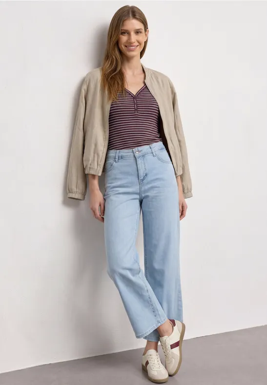 Een vrouw staat voor een witte muur, draagt een lichte rechte jeans, een gestreept shirt en een beige jasje.
