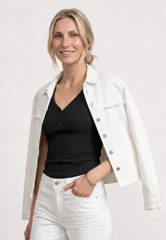Frau in schwarzem Top, weißer Jeansjacke über den Schultern und weißen Jeans vor neutralem Hintergrund.