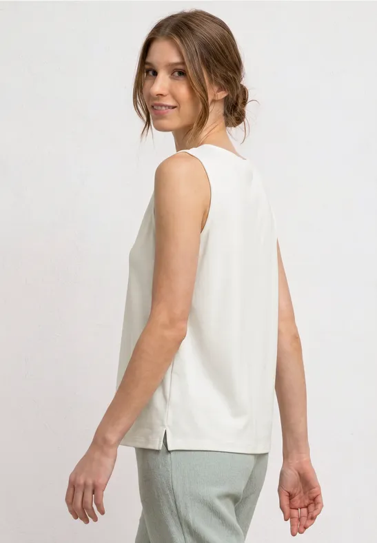 Een vrouw draagt een witte mouwloze top en grijze broek, zij-aanzicht.