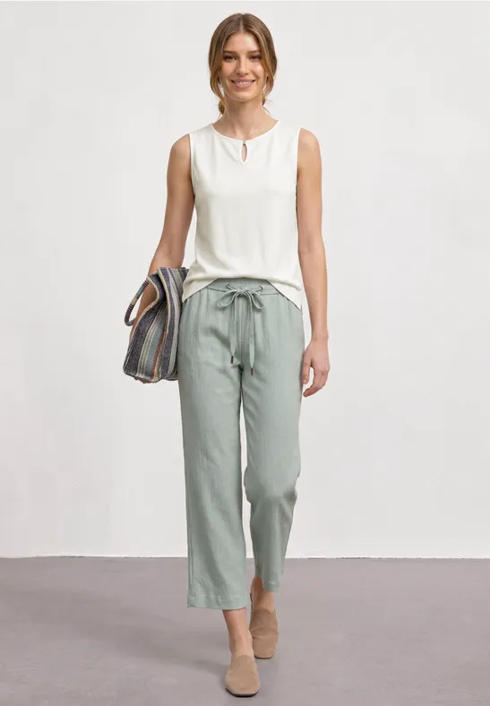 Vrouw in witte mouwloze top en lichtgroene broek met strikceintuur, beige schoenen en gestreepte tas.