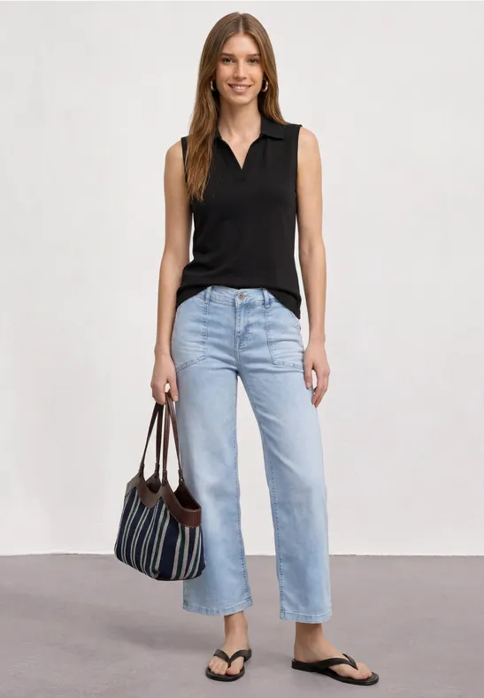 Femme portant un haut noir sans manches, jean clair, tongs noires et sac rayé sur fond neutre.