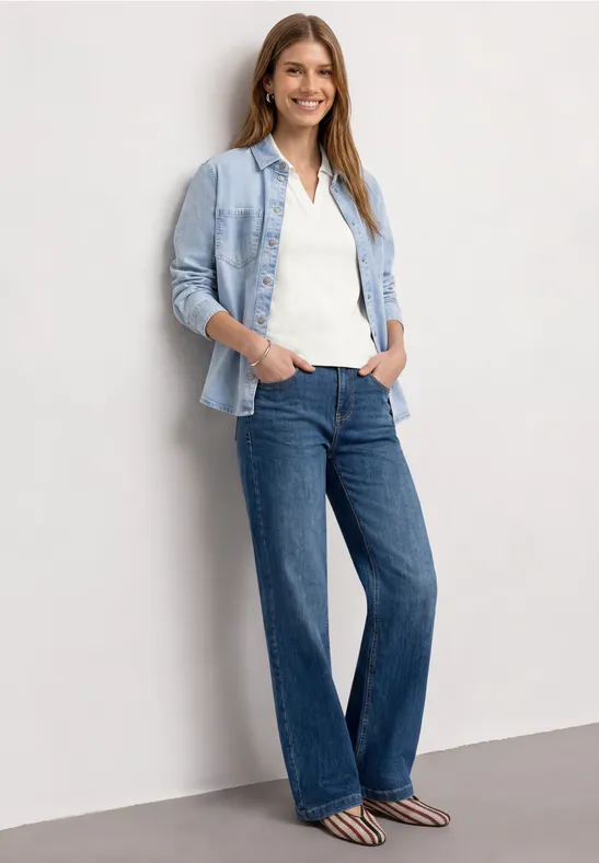 Eine Frau lehnt an einer Wand, trägt helle Bluse, Jeanshemd, weite Jeans und gestreifte Schuhe.