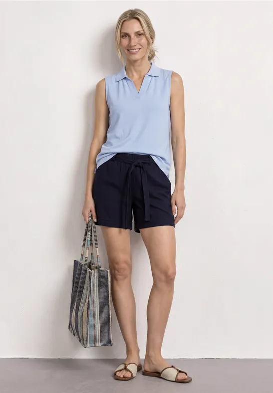 Femme portant un polo sans manches bleu clair, des shorts bleu marine, un sac rayé et des sandales sur fond blanc.