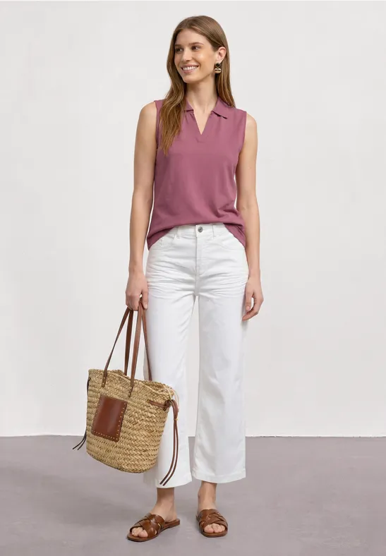 Vrouw in een mouwloze roze top, witte broek en bruine sandalen met een gevlochten strotas.
