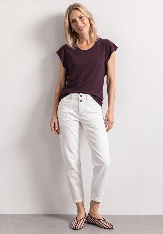 Een vrouw draagt een los T-shirt, witte broek met knopen en sandalen voor een lichte achtergrond.