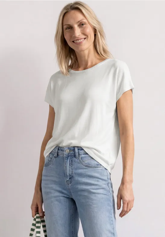 Frau in weißem T-Shirt und hellblauen Jeans vor hellem Hintergrund.