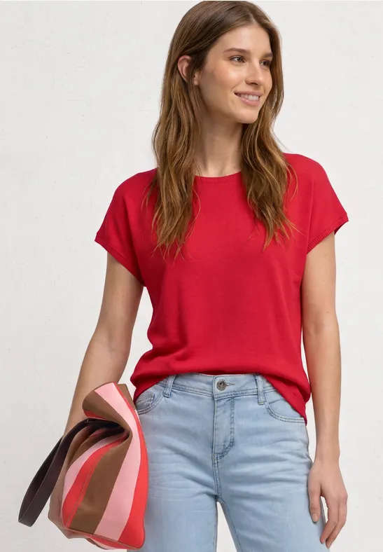 Vrouw in een rood t-shirt en lichtblauwe spijkerbroek houdt meerdere gevouwen bovenkleding vast voor een witte achtergrond.