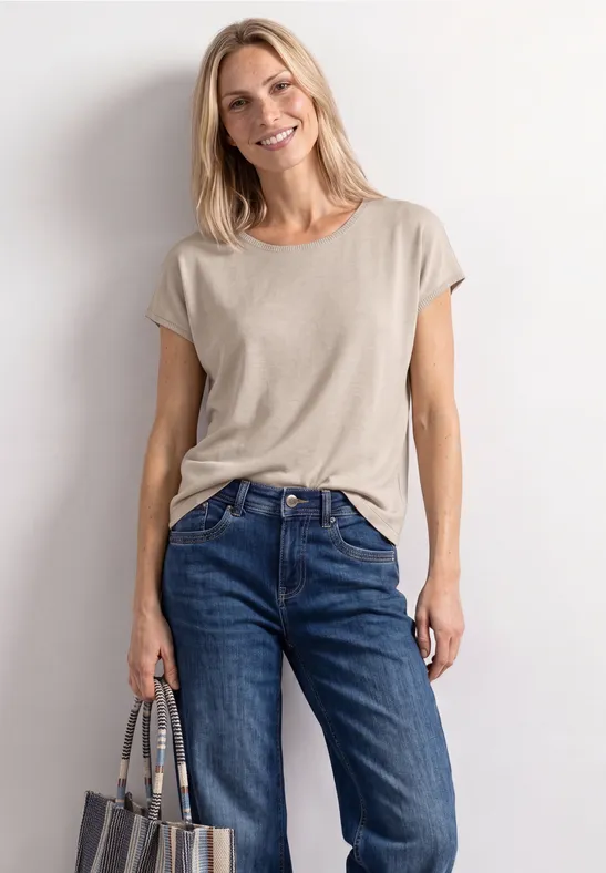 Vrouw voor een witte wand, draagt een beige T-shirt, blauwe jeans en houdt een tas in haar hand.