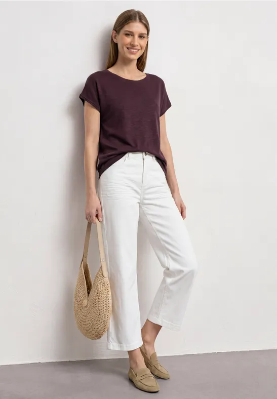 Vrouw in donkerpaars T-shirt, witte 7/8-broek, beige loafers en strozak, tegen een witte muur.