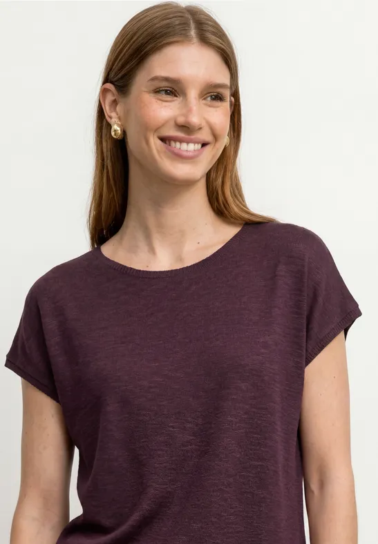 Vrouw met lang haar draagt een effen donkerpaars t-shirt en gouden oorbellen voor een witte achtergrond.