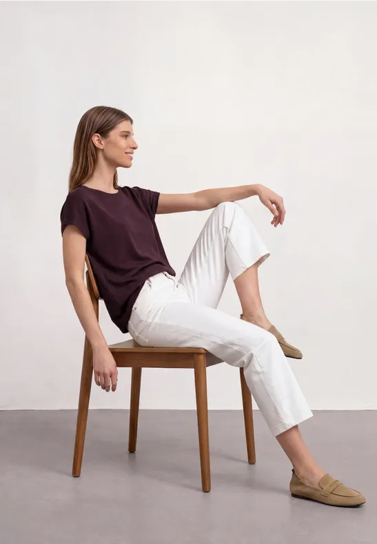 Jonge vrouw zit op een stoel, draagt een bordeaux t-shirt, witte broek en beige loafers.